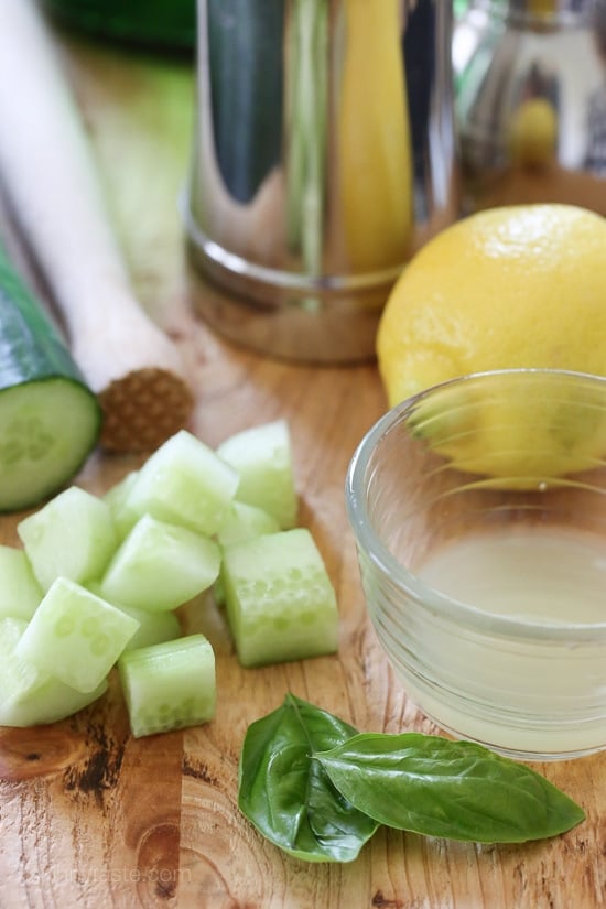 A cocktail shaker, a muddler, a whole cucumber with diced pieces, a whole lemon and a bowl of lemon juice, and basil leaves